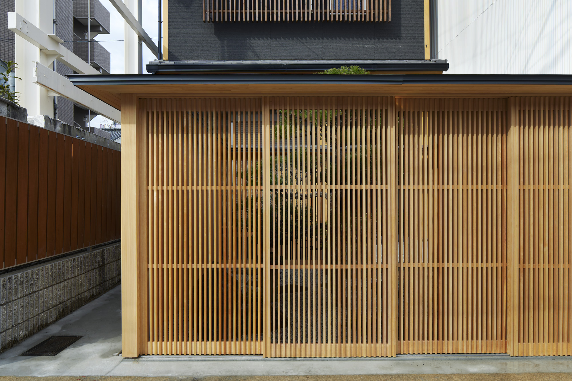 
Enclosed fence in Kyoto puts the residence’s living environment together, while serving as a gate and a backdrop of the front garden. The existing house has a frontage of 5m wide and 4.5m deep, and the space was enough for two carparking. The clients needed however no parking space, instead they would rather have a Japanese garden. They wished also to change the whole image of the existing exterior and the house by renovating this frontage into an attractive approach.

First, we planned a square-shaped enclosure in the existing open space with latticed sliding doors and gave different characters to the inside and the outside. Inside the fence you can arrange a garden like a tsubo-niwa, and the outside serves to keep the space neat and tidy; items such as bicycles, outdoor units, and cleaning tools that are necessary but not compatible with the beauty of the garden can be stored behind and they can be easily taken out for everyday use.

From this squared fence, eaves come out all around. Having the size of a roof ridge cap, the eaves keep the water out of wooden lattice, being made seamlessly of a sheet of galvanized steel. Seen from the street, the fence looks like a small building with its eaves almost resembling a roof. It softens a solitary image of the house front and creates a group of living environments in which the house, new fences, gardens, existing fences, mailbox, etc. are connected with each other. Thus, without major refurbishment of the main building itself, we could change the quality of the whole environment.

In addition to those architectural issues above, we propose a prototype as solution for the conflict between traditional cityscape of Kyoto and its current building regulations. Although Kyoto is known for its historical wooden buildings densely settled, modern urban fire prevention doesn’t allow wooden gate more than 2m high to be built. To achieve wooden space with wooden joinery, which is difficult in fire protection area, the height of the fence is kept lower than 2m, so that we could avoid installing fireproof sleeve walls additionally.

Each side of the fence has a span of 3.64 m with its height maximal 2 m, with 3/10pitched roof on top. And the wood is not treated non-combustible. In order to satisfy all of these, the columns and beams are made of steel. By using T-beam, the cross-sectional performance and shape of the beam are matched with the the roof, while the installation of the base material being kept compact and reasonable. This could be a general solution for similar conditions, as it is normally difficult to realize wooden fence with gabled roof in the urban area of Kyoto.

translated by Tota Goya
