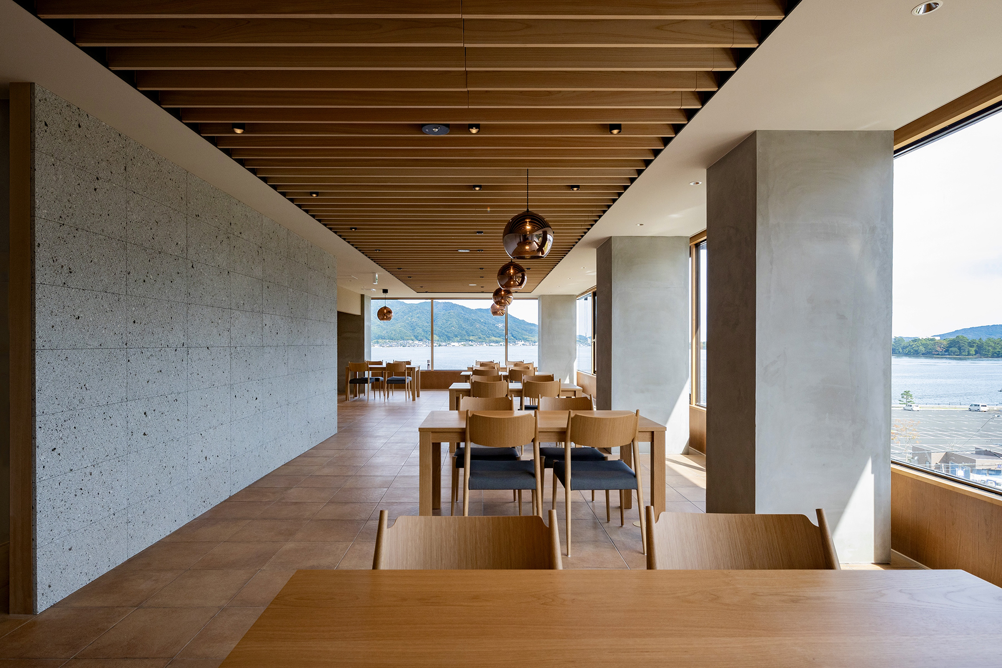 We carried out the interior renovation of the in-house restaurant at Hotel Kitanoya, located in Amanohashidate—one of Japan’s most scenic locations.
The restaurant is situated on the top floor of the hotel, offering sweeping panoramic views of Amanohashidate.
Previously used as a dining space, the area was renovated by integrating adjacent guest rooms and banquet halls, transforming it into a 300-square-meter restaurant.
To organize the irregularities of the existing floor plan, smaller spaces such as the entrance hall, private dining rooms, and back-of-house areas were strategically arranged along the existing indentations and protrusions.
Given the large seating capacity, a 40-meter-long, 1.6-meter-wide central passage was created to ensure clear lines of sight for staff and efficient food service circulation.
Excluding the private rooms, the seating area is divided into two zones: one facing directly toward Amanohashidate, and the other on the mountain side, which offers partial views.
In the Amanohashidate-facing zone, seats were spaced generously to create an open and airy atmosphere, allowing guests to fully enjoy the iconic landscape.
In contrast, the mountain-side zone was designed to be more intimate and subdued, using a system of lattice partitions to softly divide the space.
The partitions combine vertical hinoki (Japanese cypress) elements and horizontal black cherry wood, forming a woven pattern reminiscent of warp and weft.
These partitions are removable and can be taken down or reconfigured according to different spatial needs.
The original space included a distinctive quarter-spherical bay window. We responded to this by installing a corresponding form—walls and ceiling—in a mirrored position, creating a semi-spherical private dining room.
Interior finishes include Ōya stone, reddish porcelain tiles, hinoki, and black cherry wood.
Through this material palette, we aimed to create an atmosphere that is warm, clean, and quietly refined—neither overly formal nor too casual.