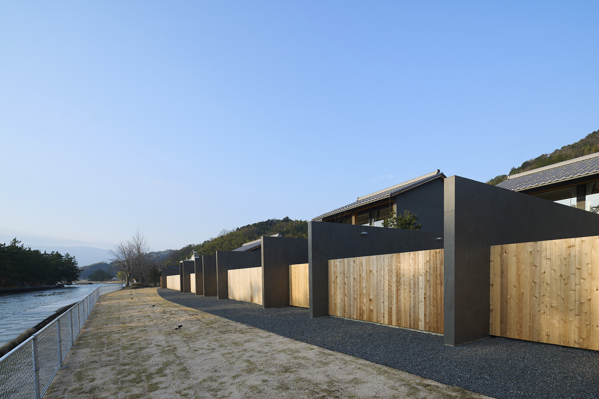 This hospitality project is located in the northern part of Kyoto Prefecture, overlooking Amanohashidate—one of Japan’s three most celebrated scenic views. The site sits along a pine-lined coastal strip, with the landscape opening toward Miyazu Bay beyond.
The project consists of four guest villas and a management building that includes a gym. Each villa is designed for private use and accommodates up to four guests, featuring a private pool, open-air bath, and sauna. While the program remains consistent, the spatial configurations differ across the four villas.
Villa A emphasizes its connection to the landscape with a wide frontage facing Amanohashidate. Villa B is arranged around a central courtyard that organizes the flow of movement. Villa C offers a linear sequence of spaces, connecting the entrance hall, wet area, and a framed view of the bay. Villa D enables direct access from a second-floor bedroom to the pool below. By varying the relationships among shared elements, the design offers a distinct experience in each villa.
The ensemble comprises two single-story and two two-story villas. Roof shapes, eave depths, heights, and building positions are calibrated to maintain visual coherence while introducing variation, allowing continuity and contrast to coexist across the site. To preserve the character of the area, roofing materials are regulated under local landscape protection guidelines. The ensemble adopts a gently sloped tiled roof, designed to form a gently articulated skyline that harmonizes quietly with the surrounding landscape.
A defining feature of the composition is a series of wall elements running orthogonal to the Amanohashidate shoreline. These walls simultaneously segment and connect the villas, introducing rhythm and order across the site. Each wall continues uninterrupted from interior to exterior, clad in the same porcelain tiles used for the outdoor flooring. The detailing throughout—walls, floors, pools, and baths—strictly adheres to the tile grid, with precise alignment. In doing so, the design avoids treating features such as pools or open-air baths as mere amenities, instead integrating them as core architectural components.
A pedestrian promenade runs alongside the waterfront, presenting a need to balance openness with privacy. To address this, the floor level of each unit is raised approximately 200mm higher than in typical timber construction, creating a subtle elevation difference. On the waterfront side, the terrace is enclosed by a parapet wall set at 1100mm high—deliberately calibrated to block sightlines from passing pedestrians. While it prevents visual intrusion from the outside, this configuration enables uninterrupted views of the pine forest and the bay beyond from within. The sectional strategy allows framed openness to coexist with enclosure, forming a calm interior atmosphere while maintaining a connection to the landscape.
By using rooflines and wall elements as unifying devices while varying the spatial sequences in each unit, the architecture supports a diversity of experiences—offering not only richness in each stay but also an invitation to return.